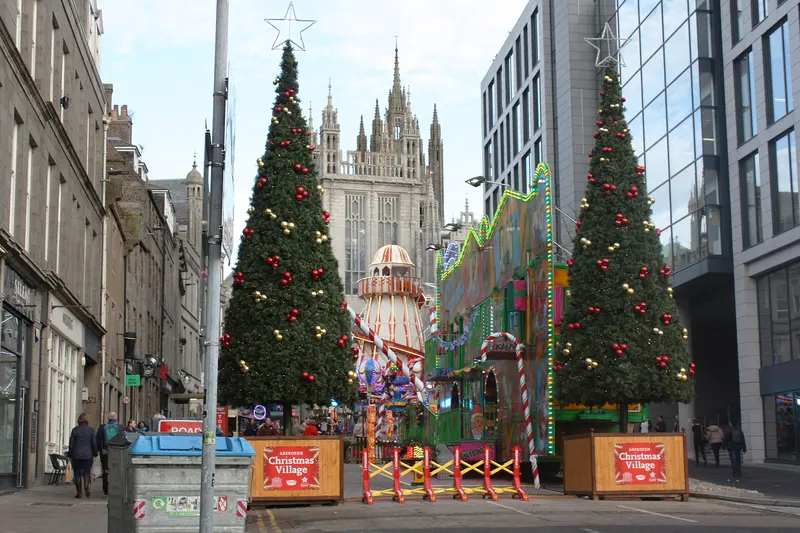 Two decorated Christmas trees and a lit-up fairground ride on Broad Street, with the granite spires of Marischal College behind