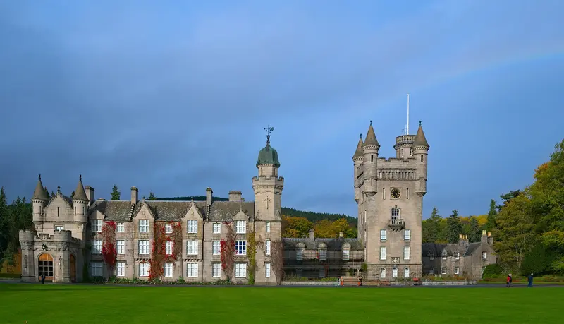 The granite towers and turrets of Balmoral Castle across a wide green lawn, with autumn trees under a grey-blue sky