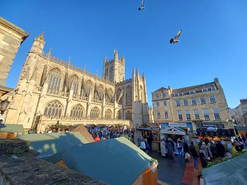 Crowds among market chalets and food stalls in front of Bath Abbey and Georgian buildings, under a clear blue sky