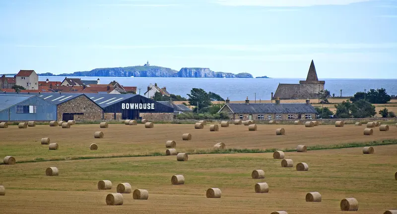 The Bowhouse farm buildings among fields of round hay bales, with St Monans church and the sea beyond