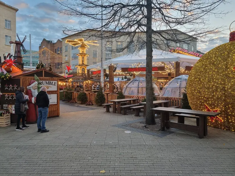Wooden market huts, picnic benches, clear dining domes, a lit Christmas pyramid and a giant golden bauble on a paved shopping street at dusk