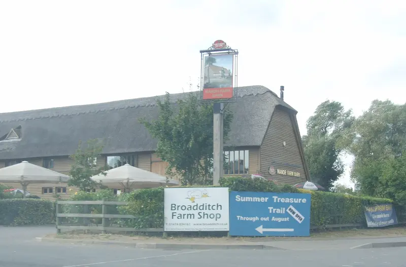 A long thatched barn behind a fence and parasols, with a hanging sign and a roadside sign for Broadditch Farm Shop