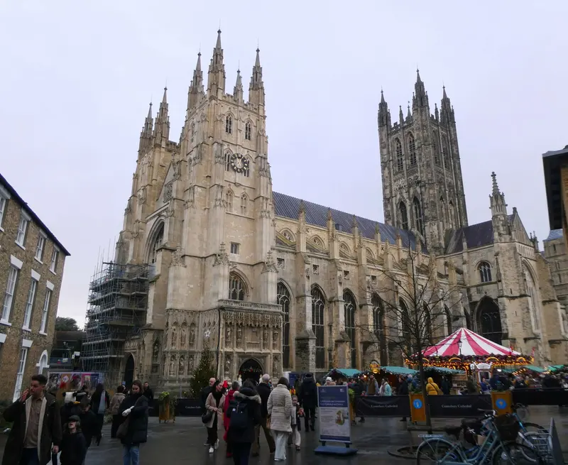 Canterbury Cathedral's pinnacled towers rising above Christmas market stalls and a red-and-white carousel, with visitors in the precincts on a grey day