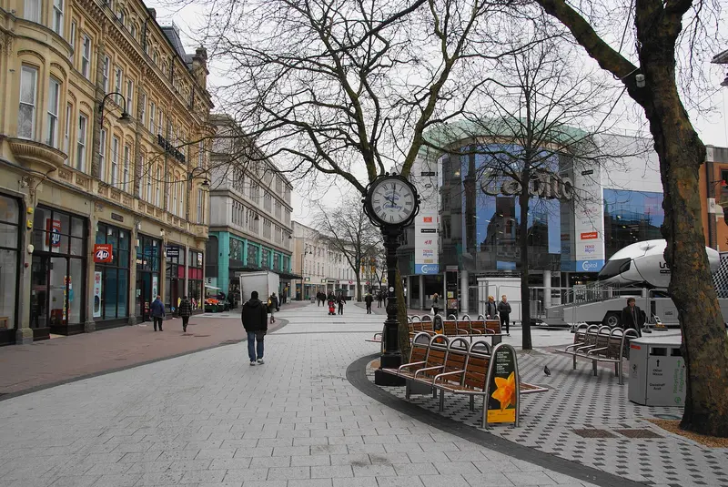 The wide pedestrianised Queen Street in Cardiff, lined with shops and trees, with a street clock and shoppers