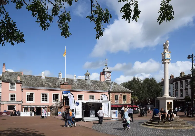 Carlisle's pink-painted Old Town Hall with its small clock turret and the tall stone Market Cross in the paved Market Square, under a blue sky with clouds