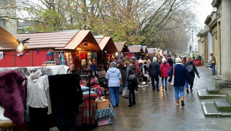 Red-roofed Christmas market chalets selling knitwear and gifts line a wet paved promenade, with shoppers walking between them under bare trees