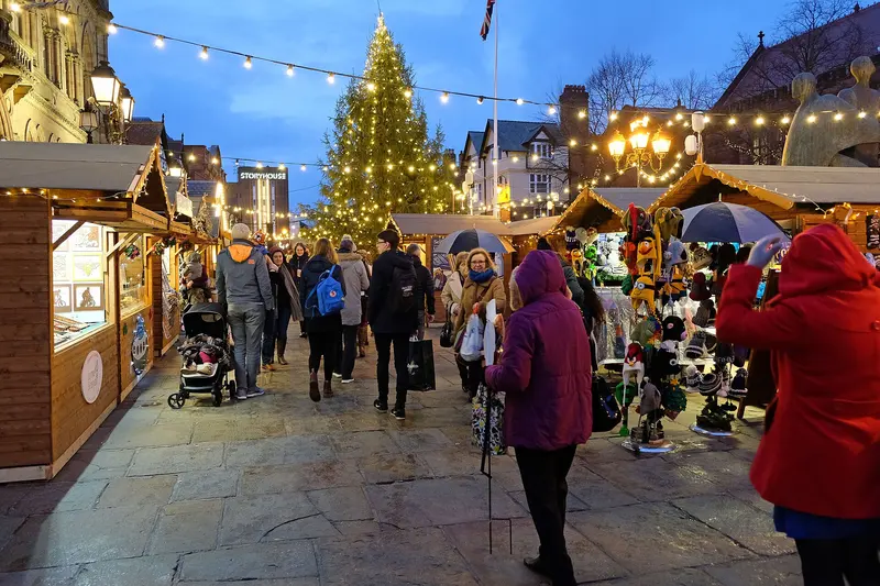 Shoppers walking between lit wooden chalets towards a tall Christmas tree covered in lights, with strings of festoon lights overhead at dusk