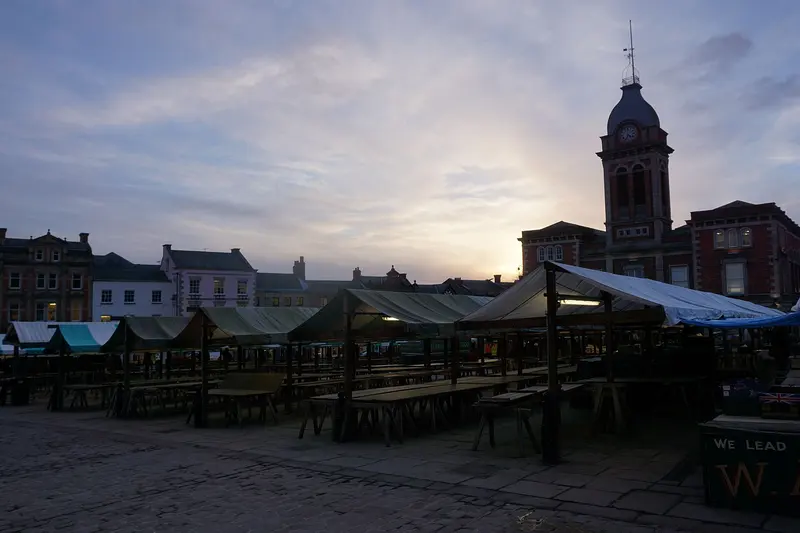 Rows of empty canvas-roofed market stalls in Chesterfield's cobbled Market Place at dusk, with the clock tower of the Market Hall silhouetted against a pale sky