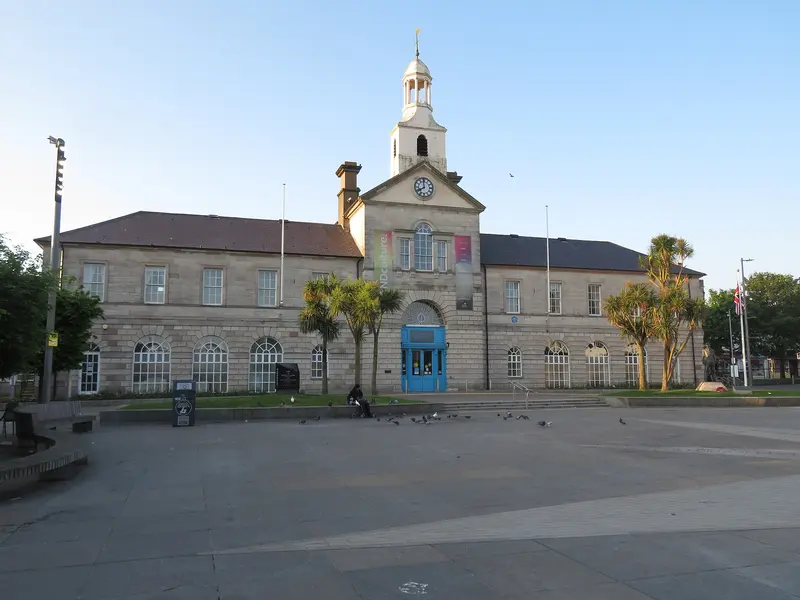 The Georgian stone Town Hall in Newtownards, with a white cupola, clock and blue doors, facing the paved Conway Square under a clear sky