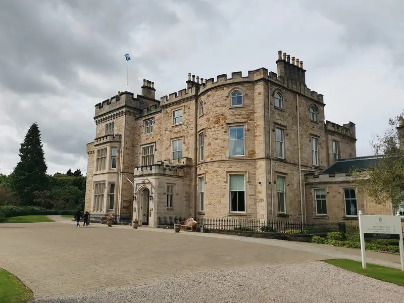 The castellated sandstone front of Crossbasket Castle, with battlements and a flag on the tower, beside a gravel drive
