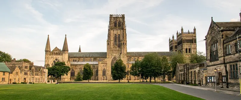 Durham Cathedral's towers and long nave across the wide lawn of Palace Green, framed by old university buildings in warm evening light