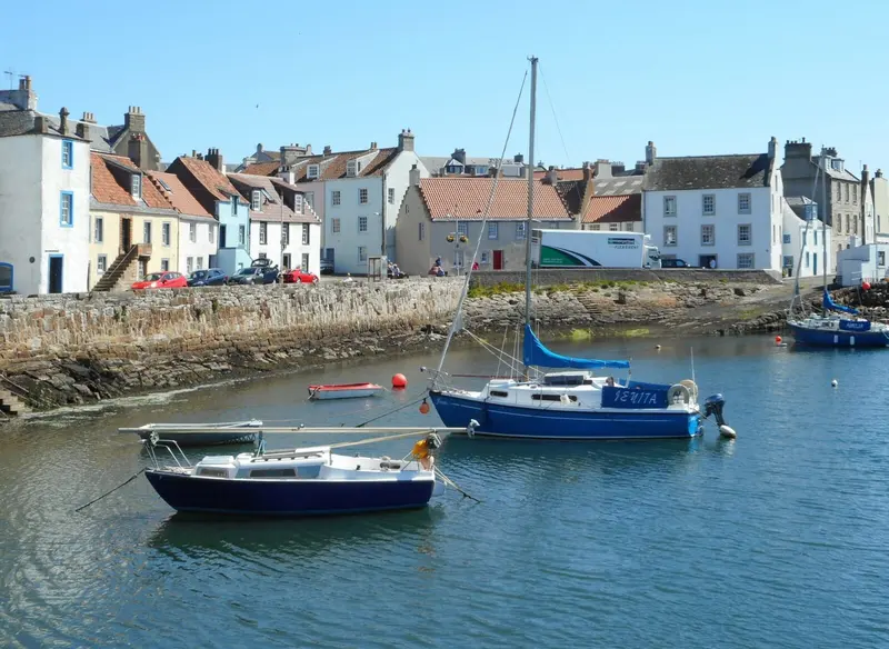 Small sailing boats moored in St Monans harbour in front of a row of whitewashed and pastel-coloured fishermen's houses under a blue sky