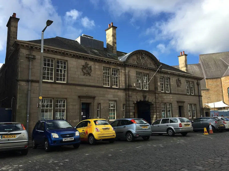 The two-storey stone front of the former drill hall on Dalmeny Street, with a carved pediment, and cars parked on the cobbles in front