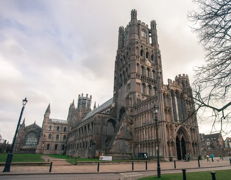 The west tower and front of Ely Cathedral rising above a lawn, with bare winter trees and a lamp post in the foreground