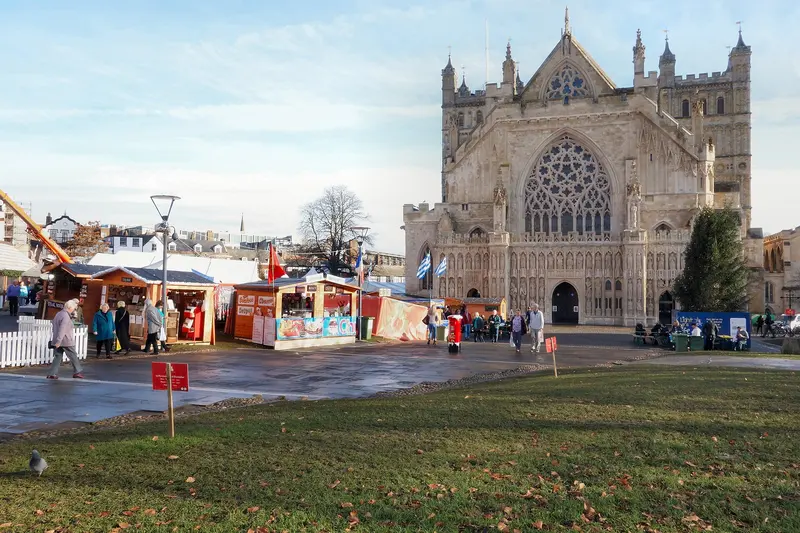 Rows of wooden Christmas market stalls on Cathedral Green, with the ornate west front of Exeter Cathedral behind and a lawn in the foreground