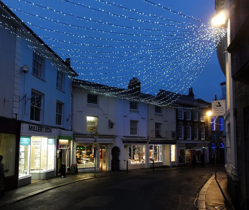 A canopy of white Christmas lights strung above a street of lit shopfronts in Falmouth town centre at dusk