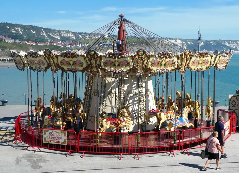 An ornate gilded carousel on Folkestone Harbour Arm, with the sea, white cliffs and the town's hillside behind under a blue sky