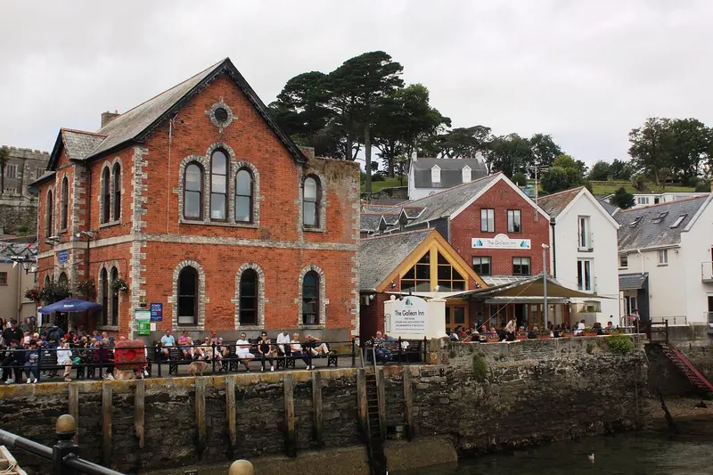 Fowey's Town Quay beside the harbour wall, with a red-brick Victorian building, painted houses and busy waterside tables