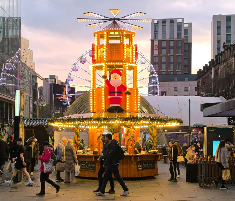 A tiered wooden Christmas pyramid bar covered in lights with a Santa figure, a big wheel behind and shoppers passing in St Enoch Square at dusk