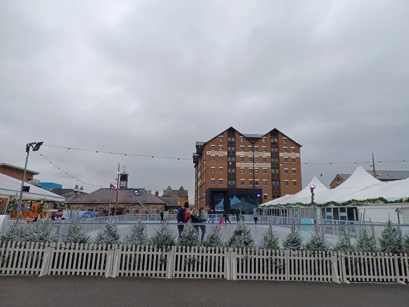 A small ice rink behind a white picket fence lined with frosted mini Christmas trees, with festoon lights, white marquees and a tall brick warehouse behind