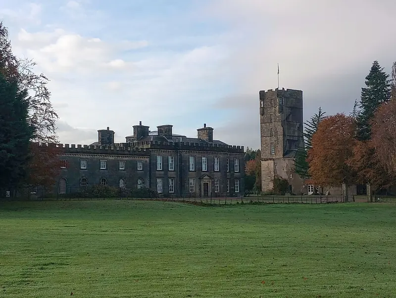 The long grey castellated front of Gordon Castle and its tall old tower beside a lawn, framed by autumn trees