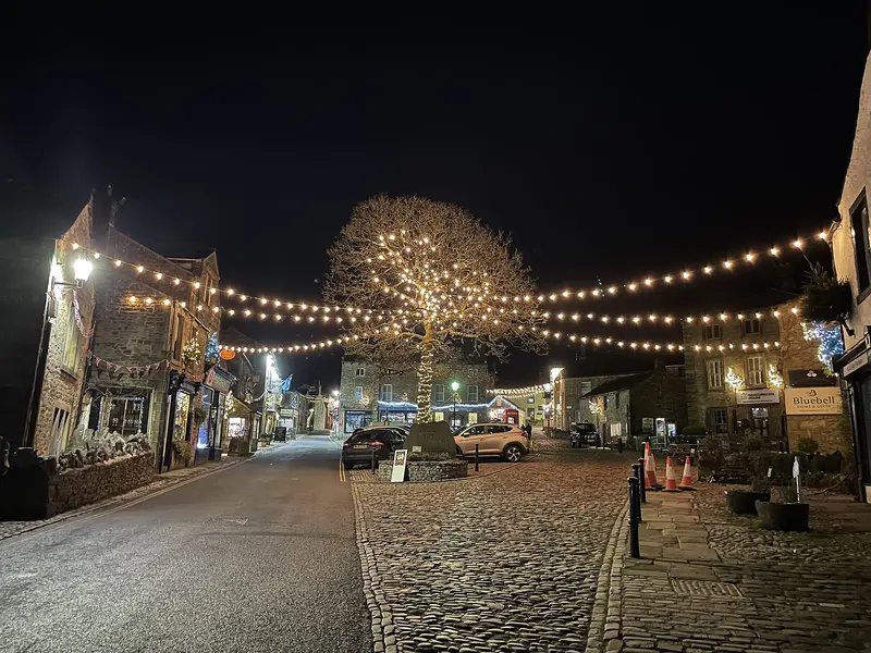 Strings of warm festoon lights and a lit tree over Grassington's cobbled square at night, with stone cottages, a Christmas tree and a red phone box