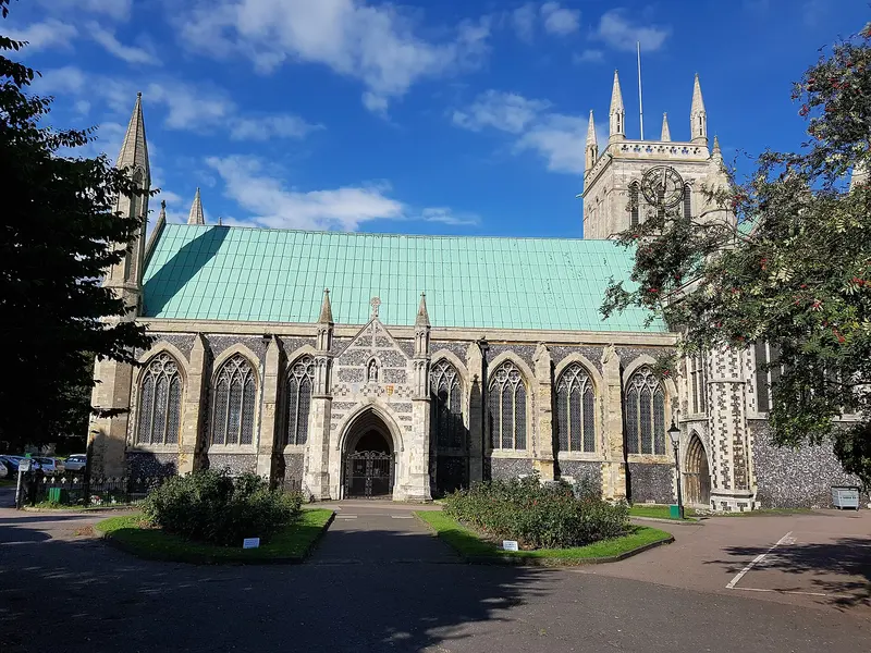 The flint-and-stone Great Yarmouth Minster, with its pale green copper roof and square tower, behind small flower beds under a blue sky