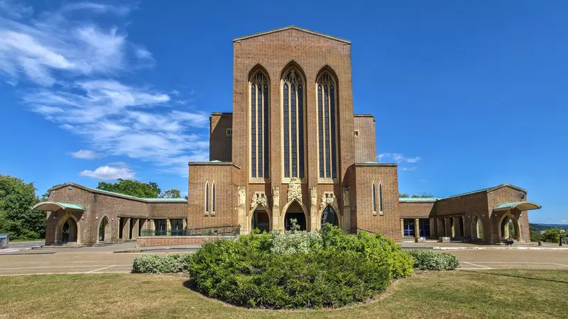 The red-brick west front of Guildford Cathedral, with three tall lancet windows, behind a round flower bed and lawn under a blue sky