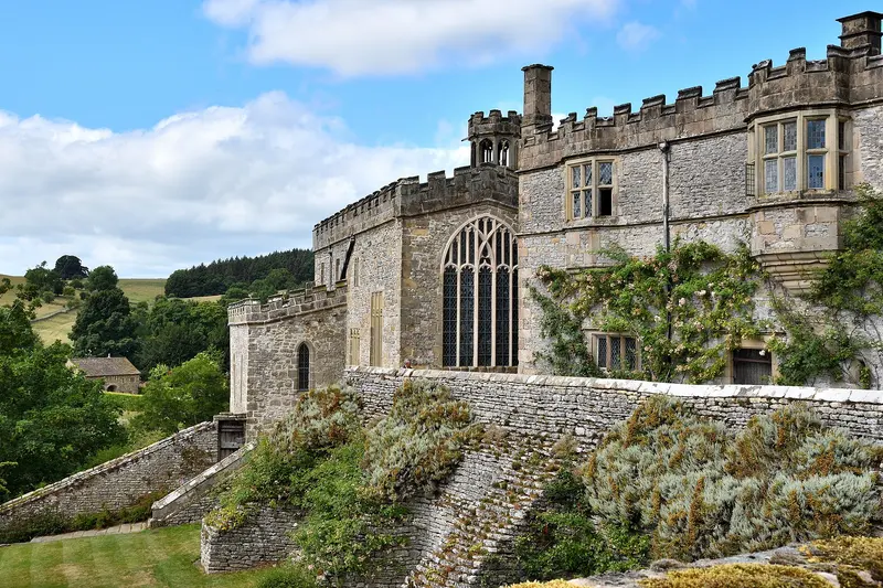 The battlemented grey stone ranges of Haddon Hall, with a large Gothic chapel window, rising above terraced gardens under a blue sky