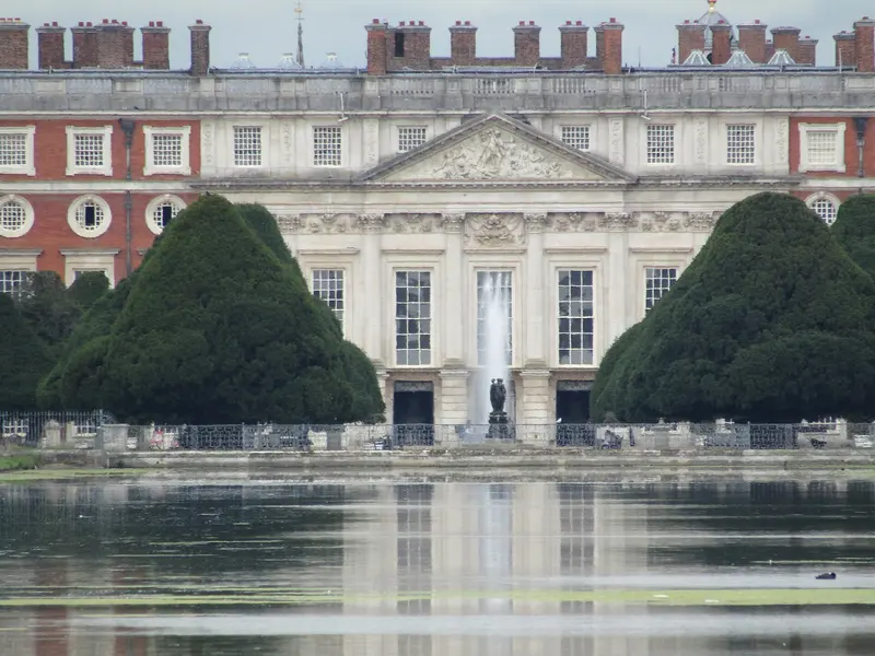 The Baroque east front of Hampton Court Palace with a fountain, framed by giant clipped yew trees and reflected in the Long Water
