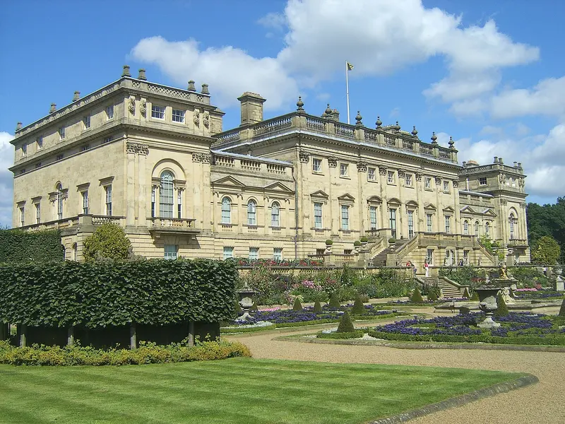 The pale stone south front of Harewood House above its formal terrace garden of clipped hedges and flower beds, under a blue sky