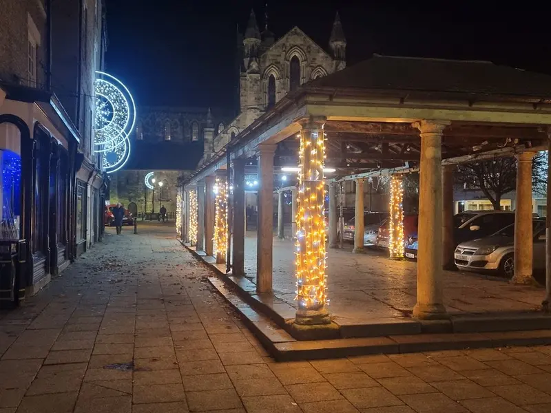 The pillars of Hexham's old covered market wrapped in warm fairy lights at night, with festive light decorations and the floodlit abbey behind