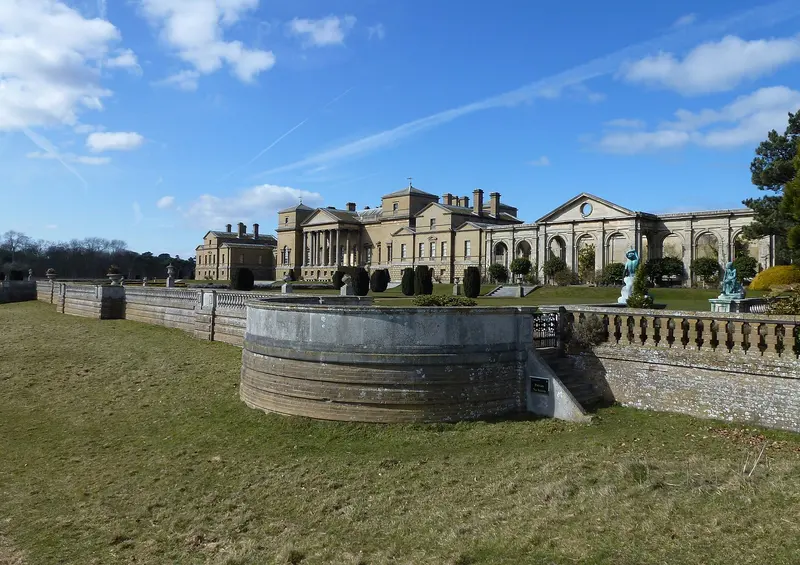 The long Palladian south front of Holkham Hall, built of pale golden brick with a columned portico, beyond a stone fountain basin and lawn