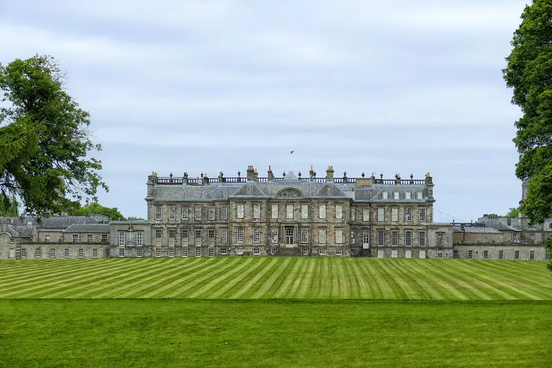 The long classical front of Hopetoun House with its central block and curved wings, beyond a wide striped lawn under a cloudy sky