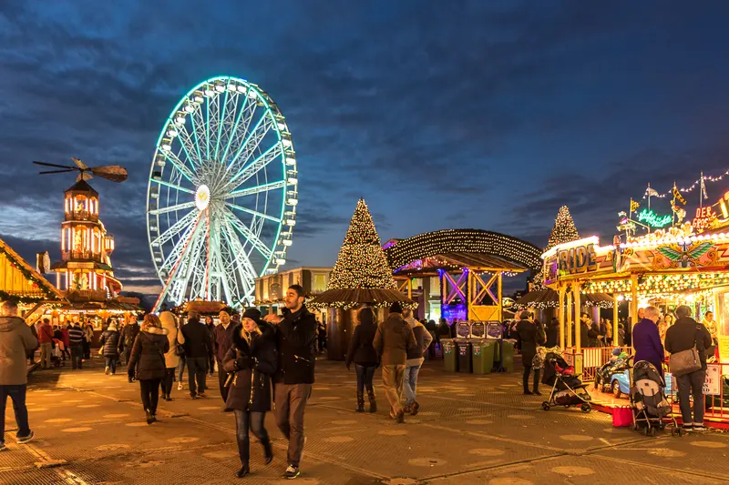 Visitors stroll past lit fairground rides, a Christmas tree of lights, a wooden Christmas pyramid and a big wheel glowing against an evening sky