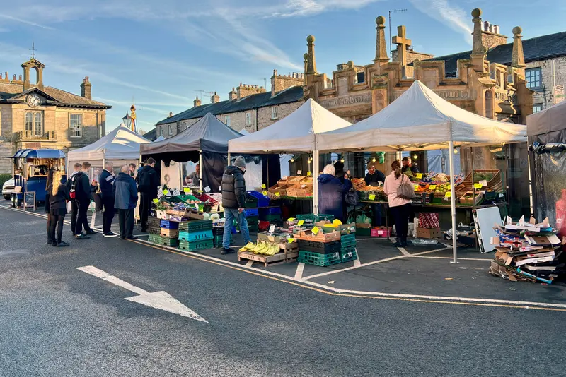 Gazebo market stalls of fruit and vegetables in Kirkby Lonsdale's Market Square in low winter sun, with the ornate stone market cross shelter and clock-towered building behind