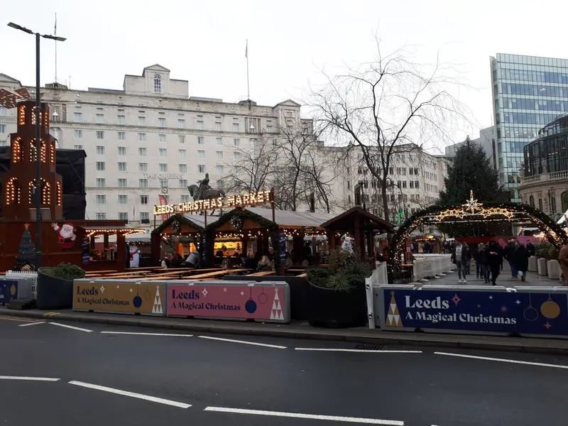 Wooden chalets, a light-up arch and a Christmas pyramid behind festive barriers in City Square, Leeds, with the white Queens Hotel behind