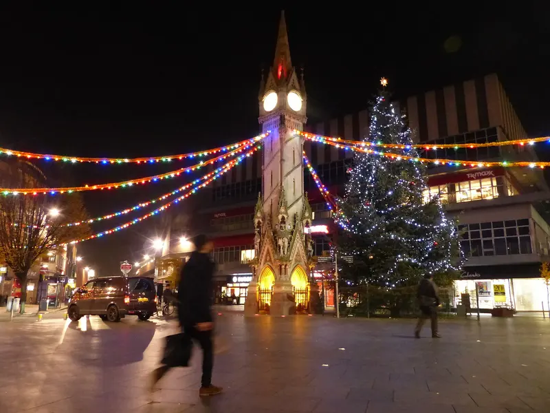 Leicester's Gothic Haymarket Memorial Clock Tower at night, strung with coloured festoon lights beside a tall lit Christmas tree