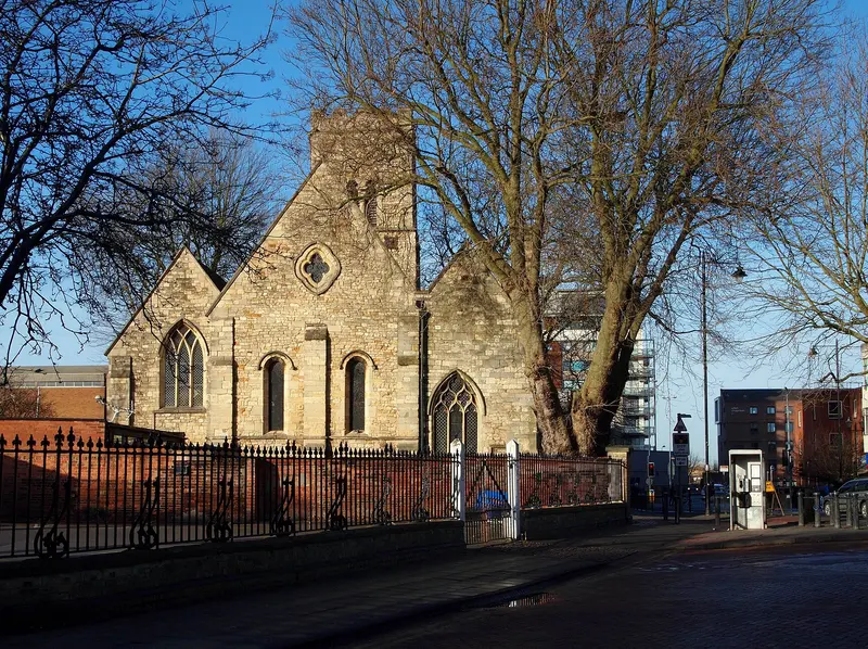 The pale stone medieval church of St Mary le Wigford behind iron railings and a bare tree, lit by low sun against a blue sky