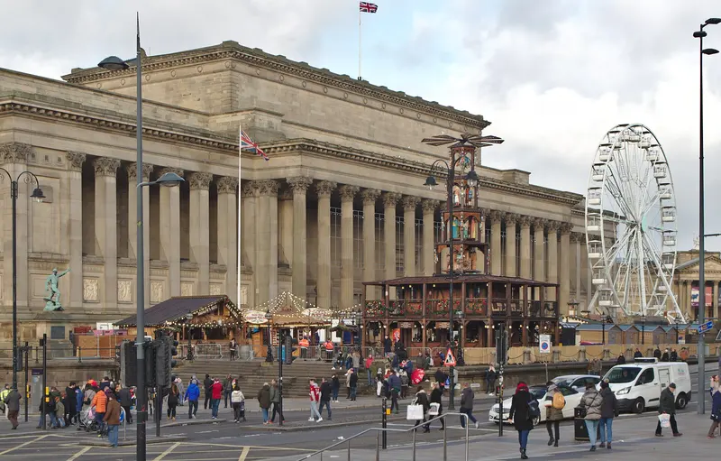 Market chalets, a tall wooden Christmas pyramid and a big wheel on St George's Plateau in front of the columned St George's Hall, with shoppers crossing the road