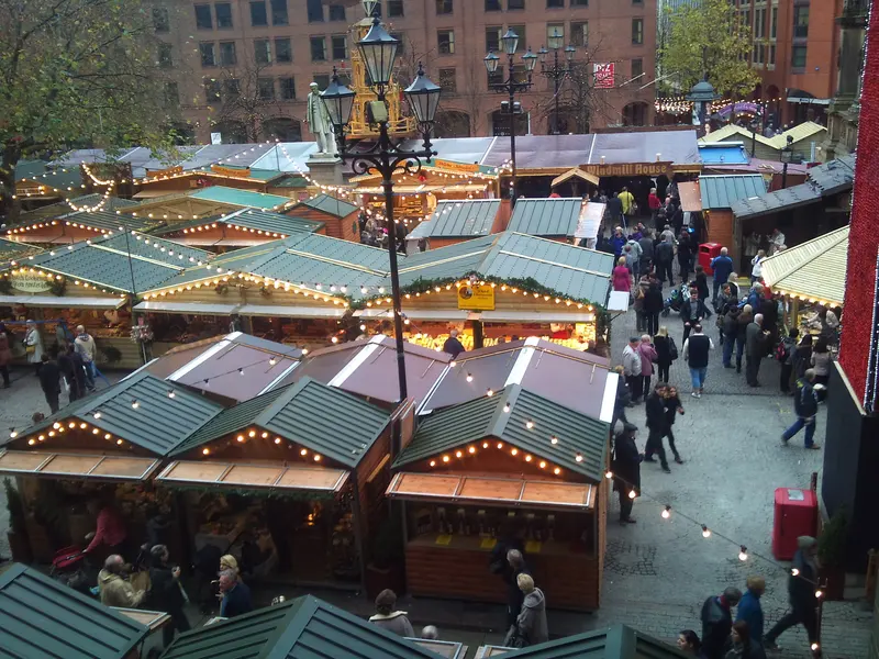 Rows of green-roofed wooden market chalets strung with lights fill Albert Square, seen from above, with shoppers walking between them