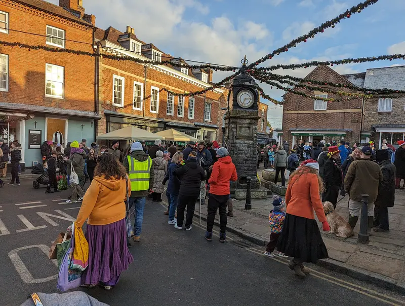 Crowds in winter coats and Santa hats gather around the clock monument in Much Wenlock's square, under festive garlands strung between brick buildings