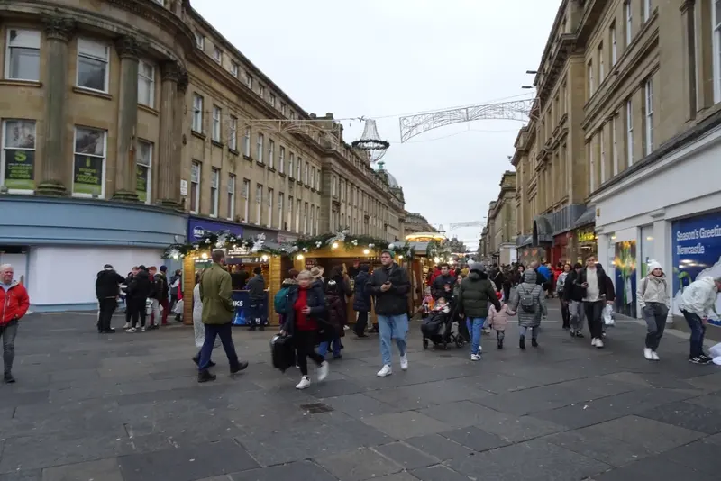 Shoppers walking between garlanded wooden market chalets on Grainger Street, Newcastle, lined with grand sandstone buildings and a light decoration overhead