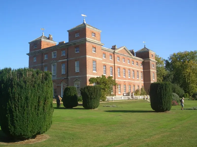 The red-brick Georgian Kimberley Hall with its four corner towers, behind a lawn and clipped yew trees under a blue sky