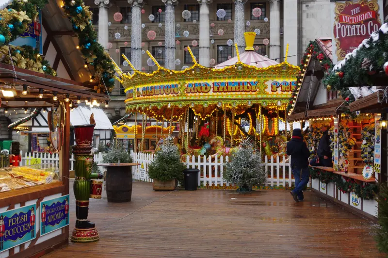 A brightly lit gallopers carousel behind a white picket fence, flanked by decorated market stalls, in front of the columned Council House