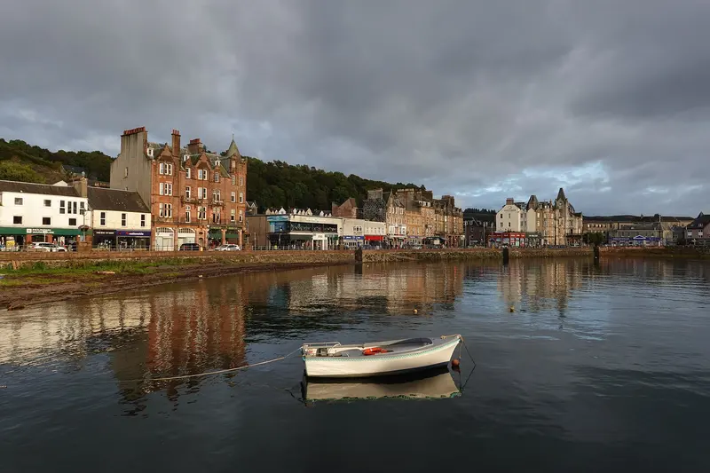 Oban's waterfront of Victorian stone hotels and shops reflected in calm harbour water, with a small white boat moored in the foreground under grey clouds