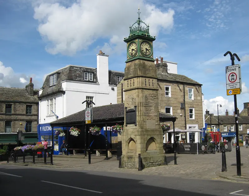 Otley's Victorian clock tower with its green copper cupola in the Market Place, beside the old covered buttercross and stone shops, under a blue sky