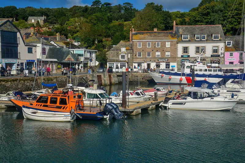 Boats moored in Padstow's inner harbour in front of a row of stone and pastel-painted houses and shops, with wooded hills behind