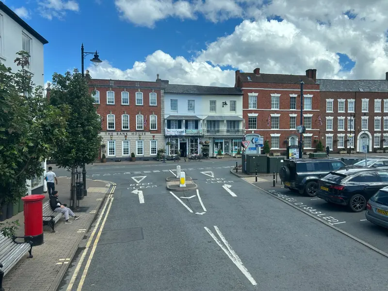 Pershore's wide Broad Street lined with Georgian brick and painted houses, with a red postbox and parked cars under a blue sky with white clouds