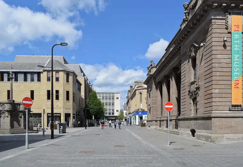Perth's pedestrianised King Edward Street, with the columned Perth Museum building on the right and the market cross and shops beyond, under a blue sky